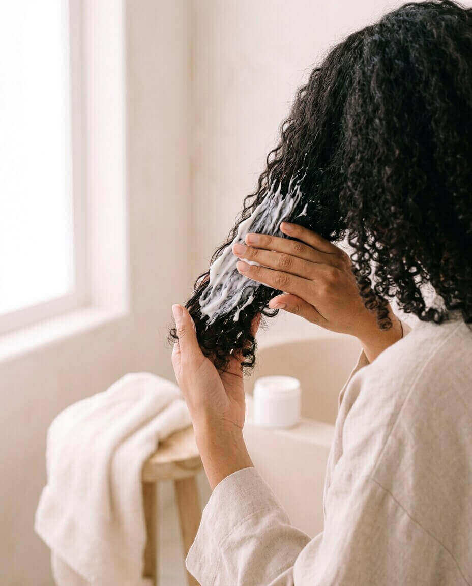 Mãos aplicando máscara em uma mecha de cabelo cacheado, vista de costas